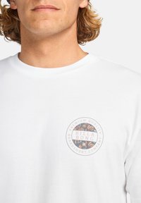 Hvit bomulls t-skjorte med et sirkulært blomstermotiv med "BILLA BONG" tekst i midten og svarte detaljer. Korte ermer og rund hals.