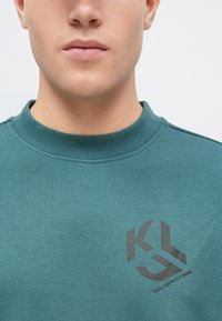 Teal sweatshirt med ribbestrikket krage, med en svart KL-logo på venstre bryst. Glatt tekstur, avslappet passform og minimalistisk design.