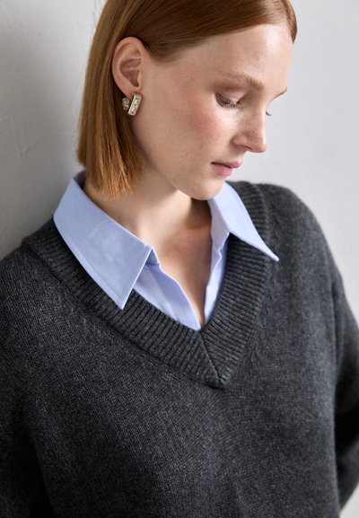 Pulover din lână gri închis, cu decolteu în V, suprapus peste o cămașă cu guler de culoare albastru deschis. Prezintă un decolteu ribbed. Accesoriul include o cercei groși cu perle.
