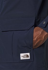 Marinblå jacka i mjukt tyg med sidfickor och en logotypmärke. Designen inkluderar en knappdetalj på ärmslutet.