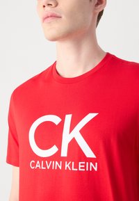Raudonas medvilnės marškinėliai su dideliu baltu "CK" logotipu ir tekstu "CALVIN KLEIN". Klasikinis apvalus iškirpimas ir trumpi rankovės, glotnus paviršius.