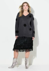 Pull en tricot gris avec surface texturée et appliqués floraux noirs, assorti d'une jupe noire frangée et de bottines métalliques.