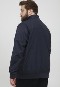 Jachetă ușoară de culoare bleumarin, cu guler stand și manșete elastice. Textură netedă, design minimal și linii de cusătură distinctive la spate.