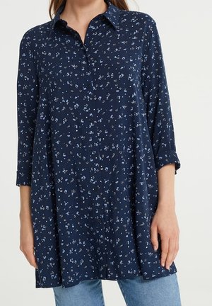 Woman wearing navy blue, long-sleeve button-up shirt with small white floral print and light blue jeans, standing against a plain background.