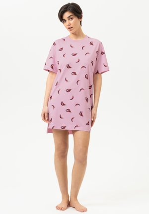Vrouw staat op blote voeten en draagt een loszittende roze jurk met korte mouwen met rode en zwarte patroon met veren op een effen achtergrond.