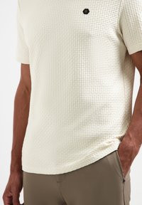 Crème-kleurig gestructureerd shirt met korte mouwen, voorzien van een geweven patroon en een klein zwart hexagonaal logo op de borst.