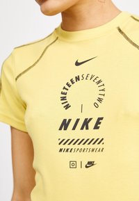 Close-up van een persoon die een gele Nike t-shirt draagt met zwarte tekst en logo's, waaronder "NINETEENSEVENTYTWO" en "NIKE SPORTSWEAR."