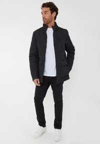 Zwarte gewatteerde jas met een hoge kraag, voorzien van een ritszak en knoopsluiting aan de voorkant, gedragen met zwarte broek en witte sneakers.