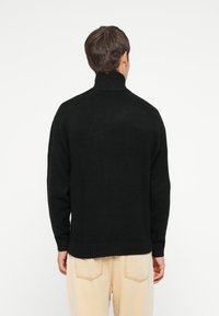 Pull en maille noir avec un col montant, des poignets et un ourlet côtelés. Le tissu texturé est présenté de dos, associé à un pantalon de couleur claire.