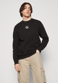 Black cotton sweatshirt with ribbed cuffs and hem, featuring a white logo on the chest. Model wears beige cargo pants.