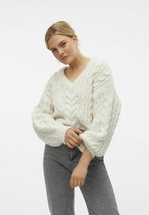 Kuscheliger weißer Strickpullover mit V-Ausschnitt, Zopfmuster und ausgeprägten Ärmelbündchen, kombiniert mit grauen hochgeschnittenen Jeans.