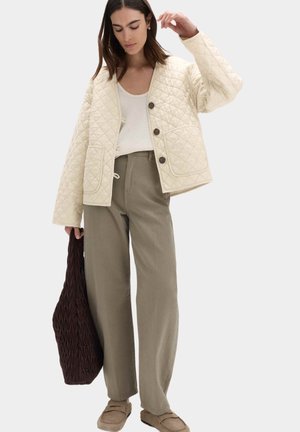 Kvinde iført cremefarvet quiltet jakke, hvid top, olivengrønne vide bukser, beige loafers, der holder en stor mørkebrun tekstureret taske.