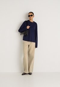 Μπλούζα με ρίγες σε ναυτικό μπλε και μωβ χρώμα, μπεζ παντελόνι με φαρδιές γραμμές, μαύρα γυαλιά ηλίου και μαύρη τσάντα. Υφασμα με υφή και άνετη εφαρμογή.