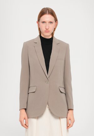 Beige skreddersydd blazer med notched lapeller, enkel knappelukking, to frontlommer og glatt tekstur. Båret over en svart topp.