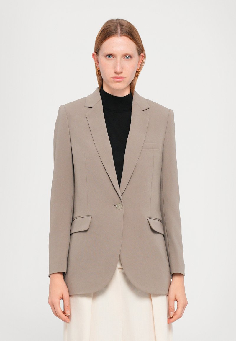 Beige skreddersydd blazer med notched lapeller, enkel knappelukking, to frontlommer og glatt tekstur. Båret over en svart topp.
