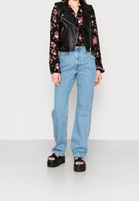 Veste de motard en cuir noir portée sur une chemise fleurie à manches longues, assortie à un jean en denim bleu clair et des sandales compensées noires.