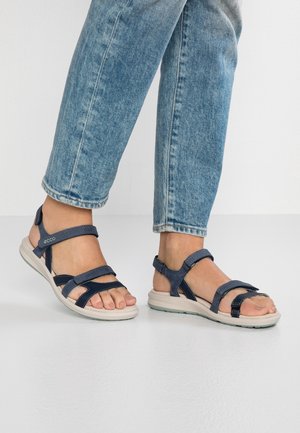 Fødder iført blå denimjeans og mørkeblå åbne sandaler med justerbare remme på en lys gulv mod en hvid væg.