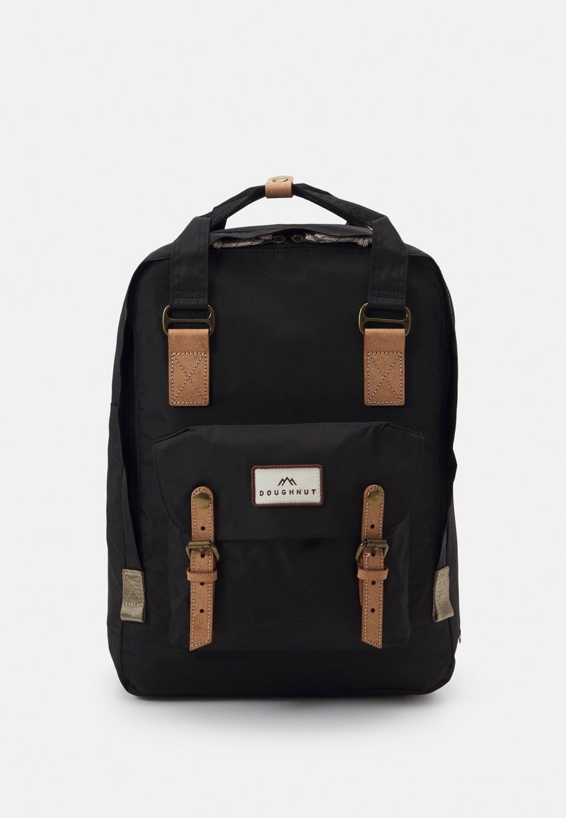 Sac à dos en tissu noir avec des sangles en cuir marron, une poche avant et une étiquette de marque. Il dispose de quincaillerie métallique et d'accents texturés.