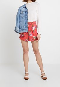 Pantaloni corti rossi con stampa floreale abbinati a una blusa bianca e a una giacca di jeans. L'outfit include sandali leggeri con tacco a blocco.