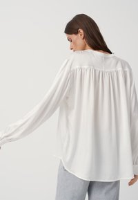 Blusa bianca a maniche lunghe con retro arricciato, texture morbida e scollatura rotonda. I polsini sono dotati di chiusura a bottone. Tessuto leggero.