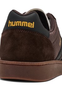 Sneaker in suede marrone con una suola in gomma testurizzata. Presenta dettagli neri e il logo "hummel" dorato sul tallone. Design semplice e profilo basso.