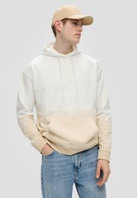 Sudadera con degradado en crema y beige, con bolsillo delantero, cordones y puños de canalé; combinada con jeans de mezclilla azul claro y una gorra beige.