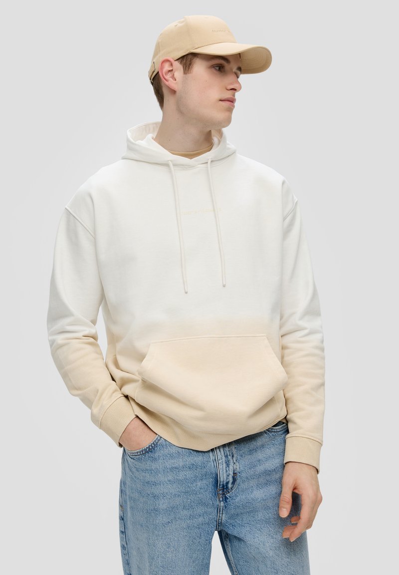 Sudadera con degradado en crema y beige, con bolsillo delantero, cordones y puños de canalé; combinada con jeans de mezclilla azul claro y una gorra beige.