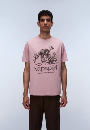Man draagt een roze Napapijri T-shirt met een afbeelding van een beer op een fiets en een bruine broek tegen een effen lichte achtergrond.