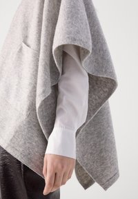 Grå uld poncho med en blød tekstur, der har et løst design og en frontlomme. Hvide skjorteærmer synlige underneath.