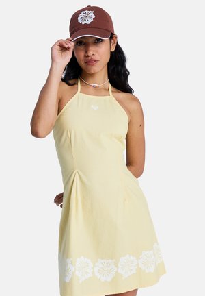 Femme portant une robe dos nu jaune pâle avec des détails floraux blancs à l'ourlet et une casquette marron avec un motif de fleur blanche.