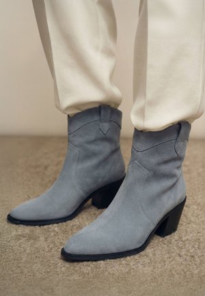 Hellblaue Wildlederstiefel bis zur Mitte der Wade mit spitzen Zehen und Blockabsätzen, getragen zu elfenbeinfarbenen Hosen und stehend auf einem teppichbedeckten Boden.