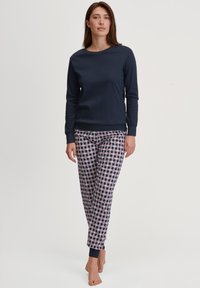 Vrouw met lang bruin haar, gekleed in een marineblauw langslippentop en gestreepte pyjamabroek, staande op blote voeten tegen een witte achtergrond.