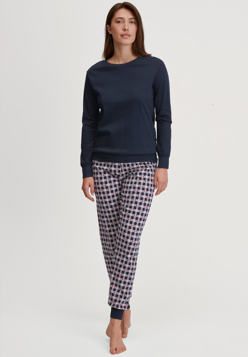 Vrouw met lang bruin haar, gekleed in een marineblauw langslippentop en gestreepte pyjamabroek, staande op blote voeten tegen een witte achtergrond.