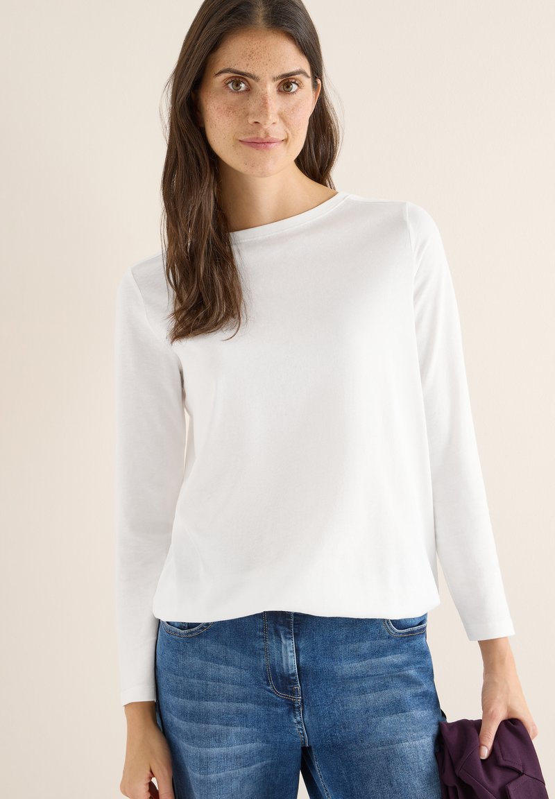 Haut blanc à manches longues avec un col rond, en tissu doux. Associé à un jean bleu et tenant un objet violet dans la main.