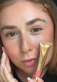 Lipgloss i en gulltube med rosa detaljer, holdt ved siden av et ansikt med klar hud, naturlig sminke og definerte øyenbryn.