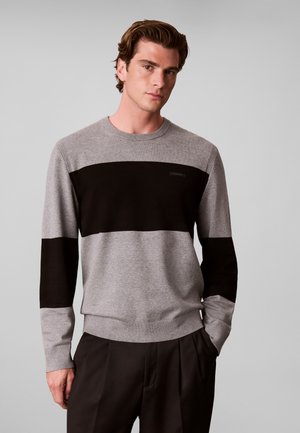 Jeune homme portant un pull gris avec de larges rayures noires horizontales sur la poitrine et les manches, associé à un pantalon noir.