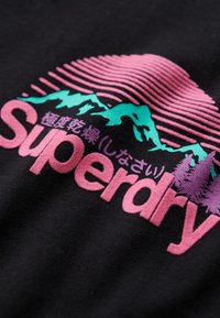Černá látka s živým grafickým designem. Růžový text "Superdry" s zelenými siluetami hor a vodorovnými linkami v odstínech růžové.