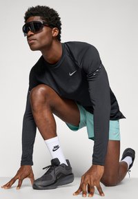 Nike langærmet sort top, mintgrønne shorts, sorte solbriller, sorte sneakers og hvide sokker med Nike-logoet, poseret i en crouched position.