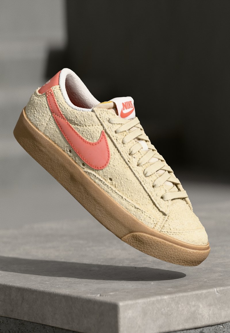 Nike sneaker in light yellow with a textured surface, pink swoosh, brown rubber sole, and white accents. Lace-up design.