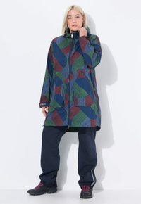 Žena v dlouhém kapucínovém kabátu s mnohobarevným geometrickým vzorem, v tmavě modrých kalhotách a černých turistických botách, stojí na bílém pozadí.