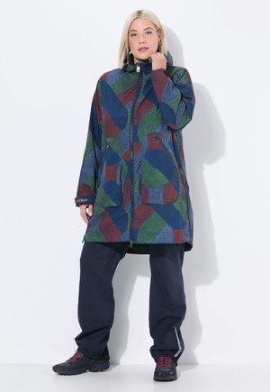 Femme portant un manteau long à capuche à motifs géométriques multicolores, avec un pantalon bleu marine et des chaussures de randonnée noires, se tenant devant un fond blanc.