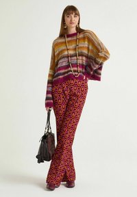 Pull en maille rayée multicolore, coupe ample, avec des accents violets ; associé à un pantalon évasé à motifs comprenant de grandes formes géométriques en rose et orange.