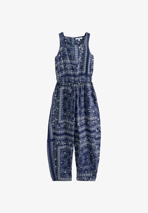Femme portant une combinaison sans manches bleue à motifs et des lunettes de soleil, debout pieds nus sur un quai en bois avec de gros rochers et un ciel dégagé en arrière-plan.