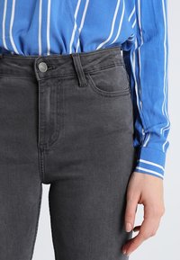 Jeans de tiro alto, de color gris oscuro con una textura suave, que presentan un cierre de botón en la parte delantera y bolsillos laterales. Combinados con una blusa de rayas azules.