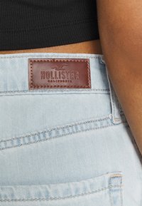 Jeans de denim azul claro com um patch de couro castanho estampado com "Hollister" e um logótipo de gaivota, com detalhes de costura visíveis.