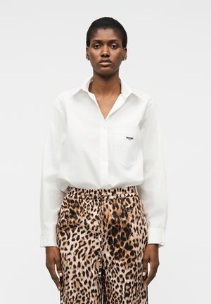 Naine, kes kannab valget nööpidega särkriietust kõrge vöökohaga leopardimustriliste pükstega, seisab otse ja tal on neutraalne väljendus.