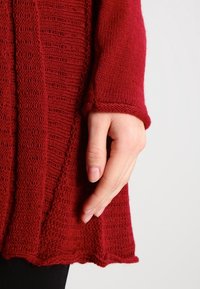 Roter Strickpullover mit strukturiertem Muster, ausgestattet mit langen Ärmeln und einem gewellten Saum. Hand in der Nähe des Saums platziert, um Maßstab zu geben.