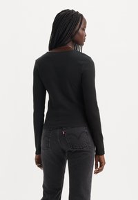 Haut noir à manches longues en côte avec une coupe décontractée, associé à un jean sombre présentant une étiquette rouge distinctive et des coutures courbes sur les poches.