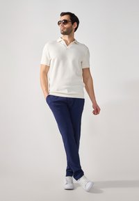 Weißes geripptes Poloshirt, marinefarbene Hose und weiße Sneakers. Der Mann trägt braune Sonnenbrillen und steht mit einer Hand in seiner Tasche.
