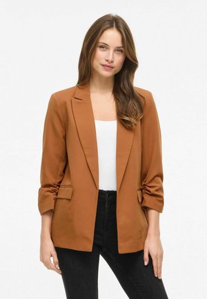 Kobieta o długich brązowych włosach, ubrana w rdzawe marynarkę na białą bluzkę i czarne spodnie, stojąca na tle białego, jednolitego tła.